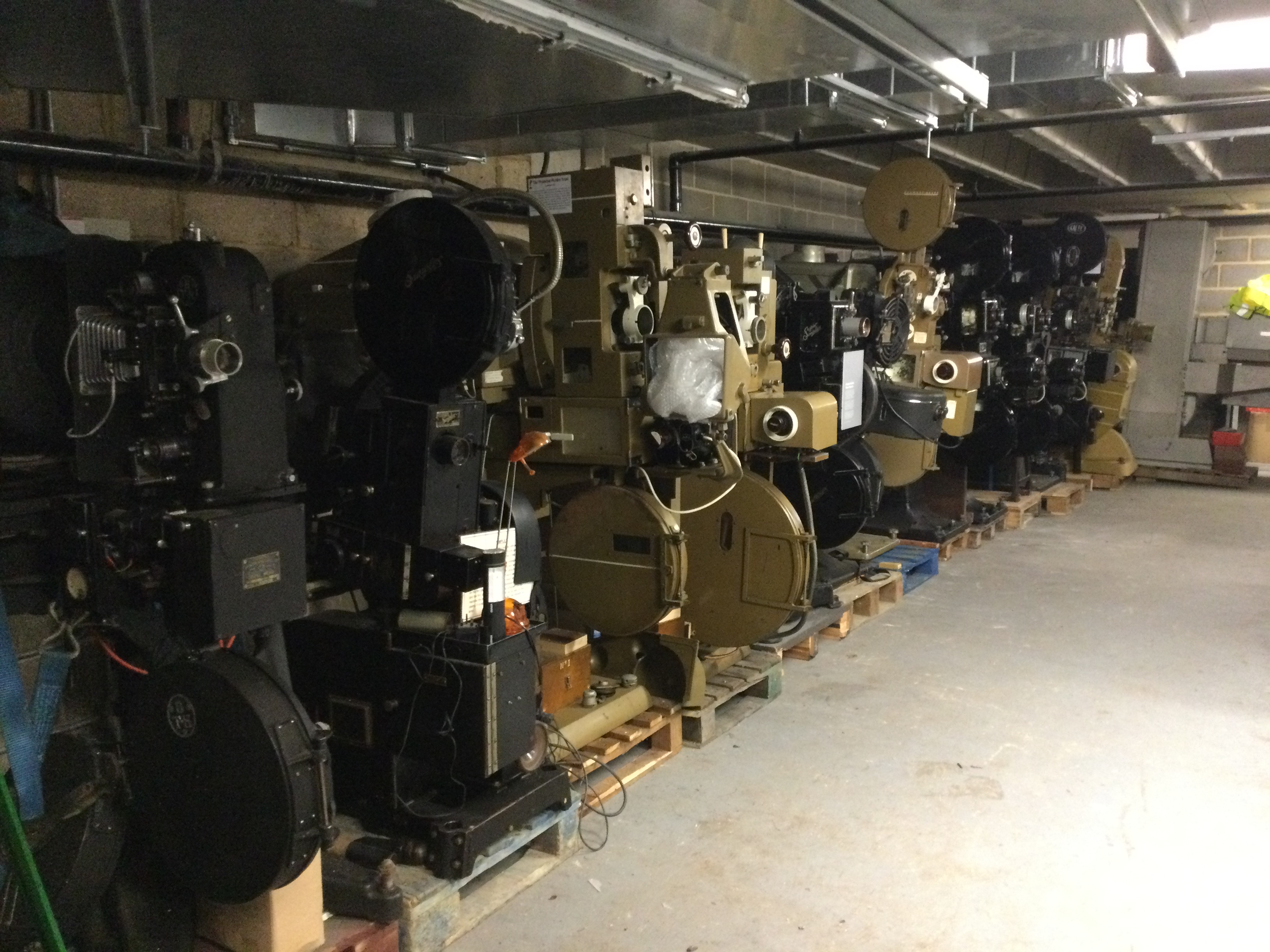 A basement storage area, with cinema film projectors on pallets lining up one side of the wall. The projectors are black and brown.