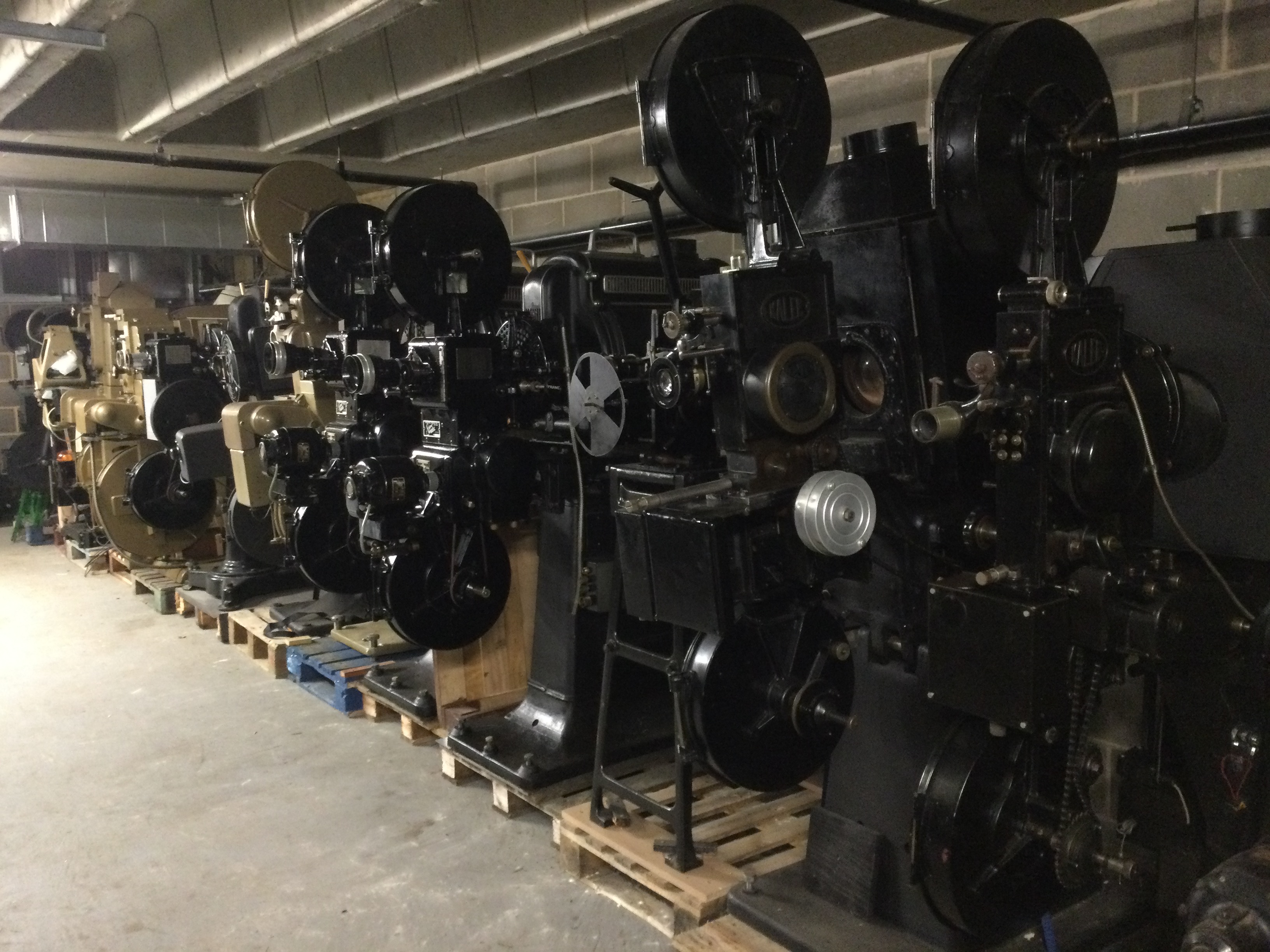 A basement storage area, with cinema film projectors on pallets lining up one side of the wall. The projectors are black and brown.