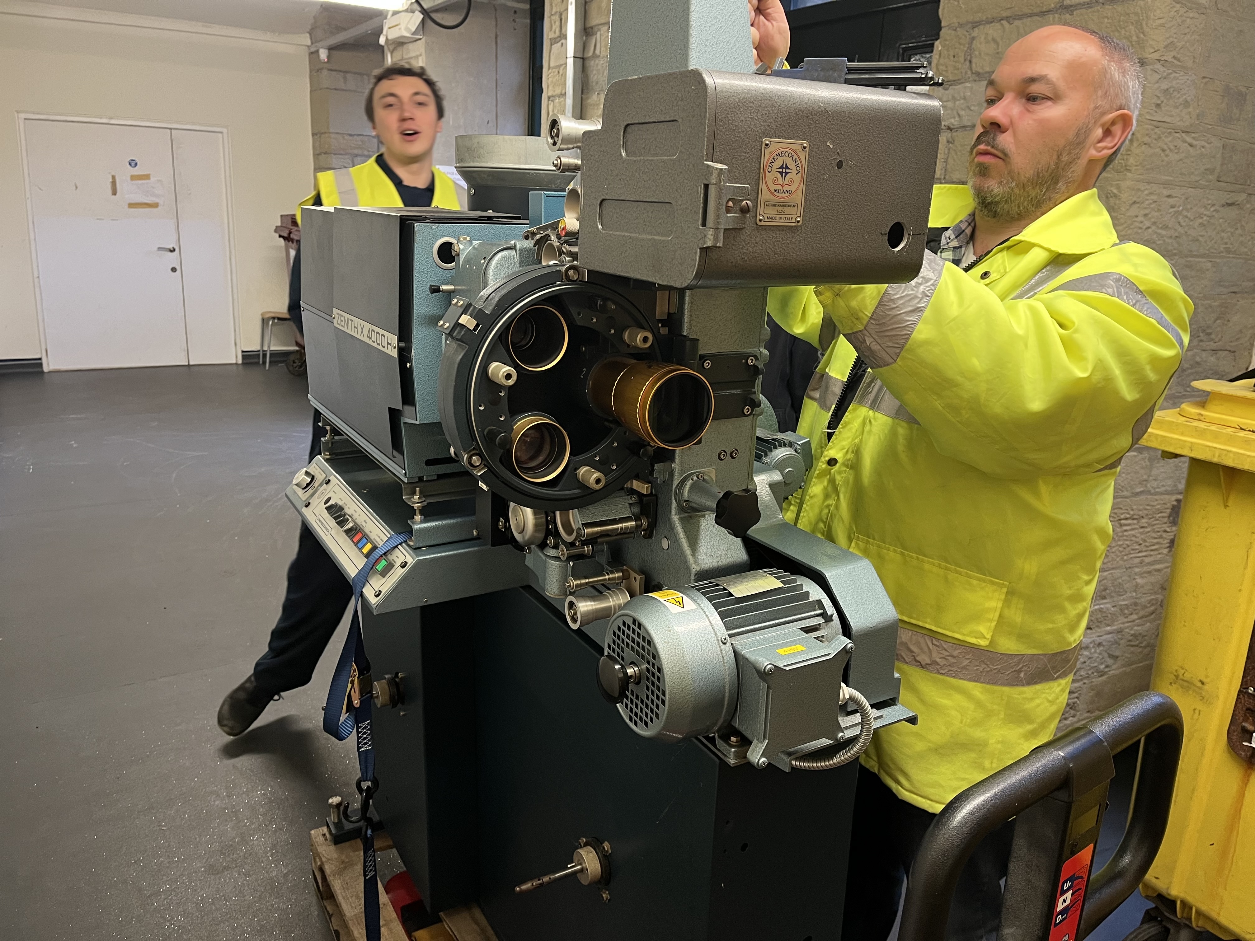 Two men wearing high visibility jackets, moving a cinema film projector. The man on the right is unscrewing a part to make it fit through the door.