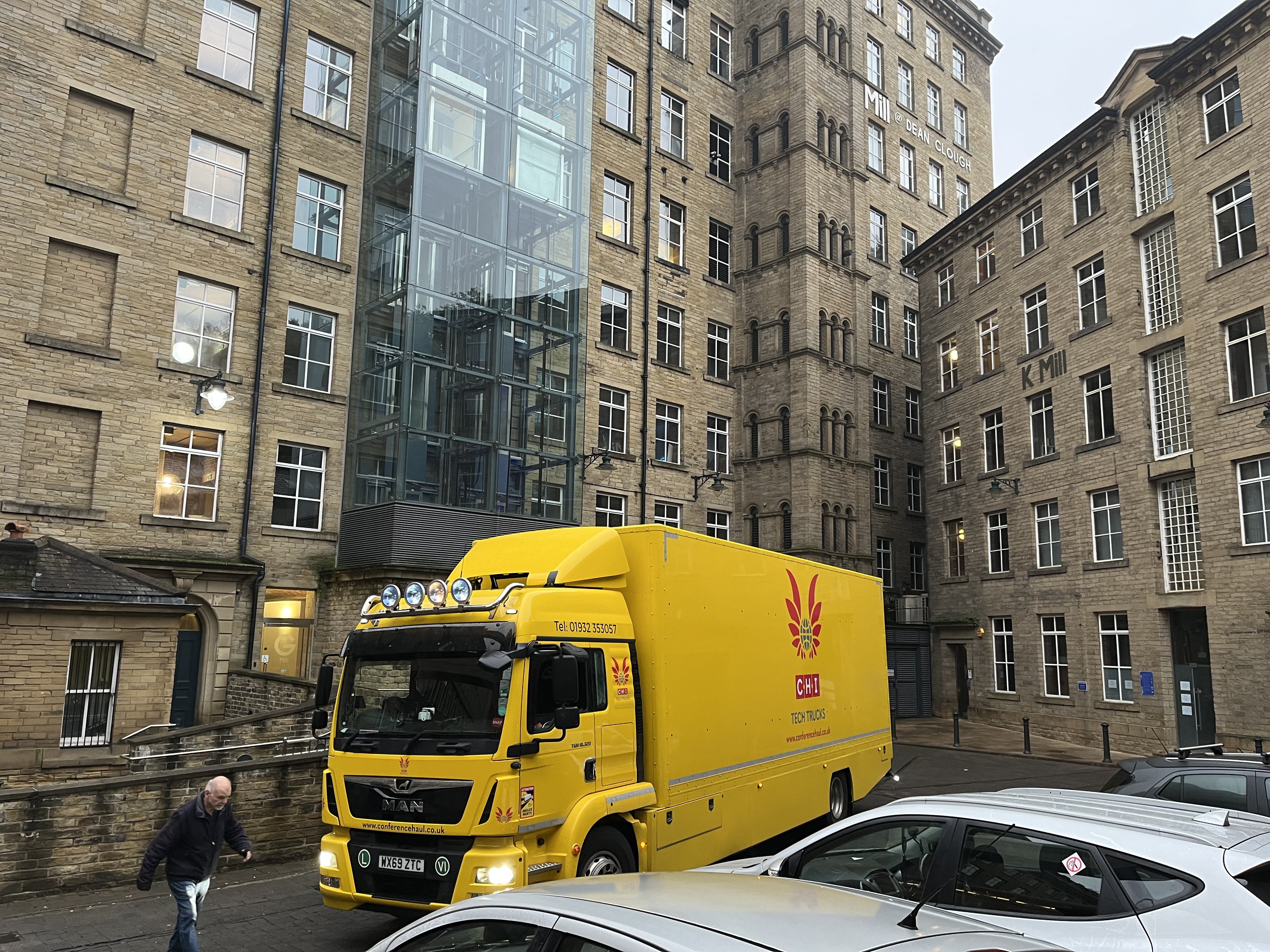 A yellow lorry arriving at an old factory building. A man walks in front of the lorry guiding it in.