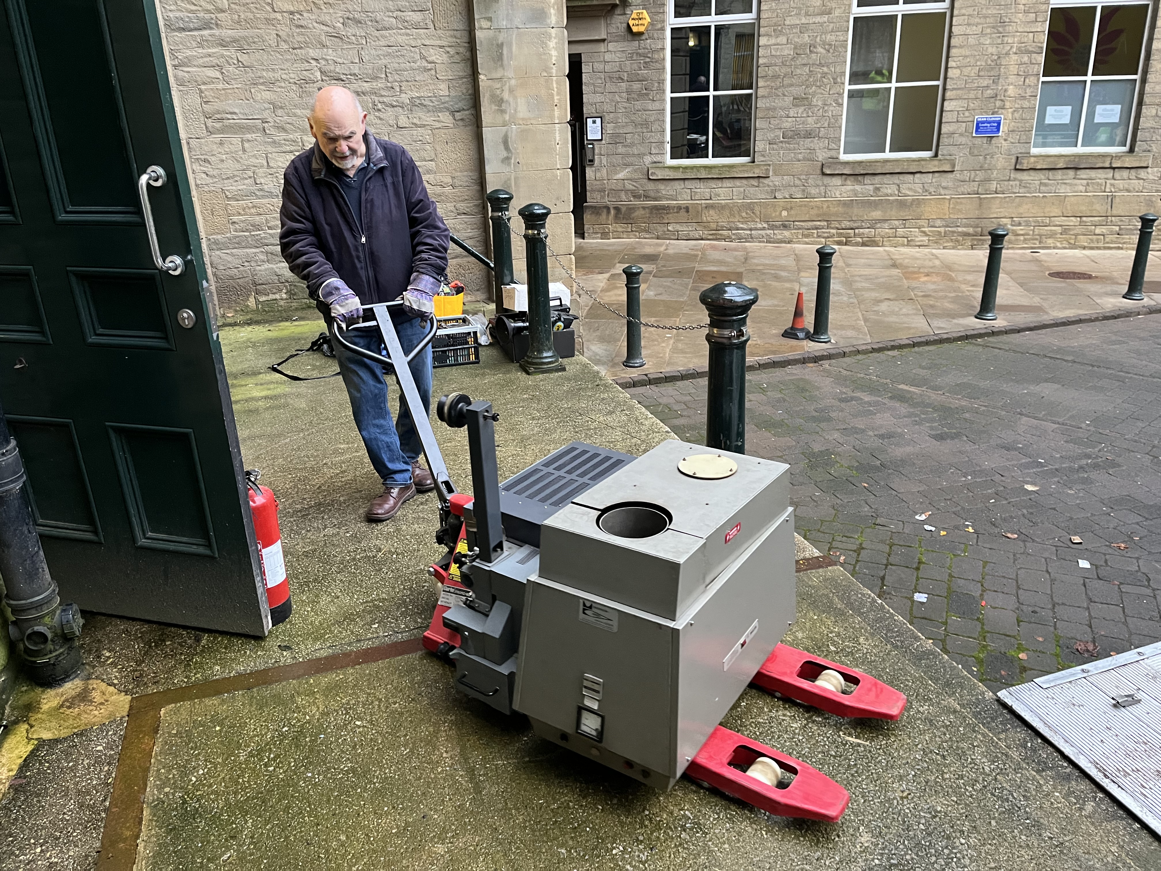 A man uses a pallet truck to move a gray rectangular lamphouse and other film projector.