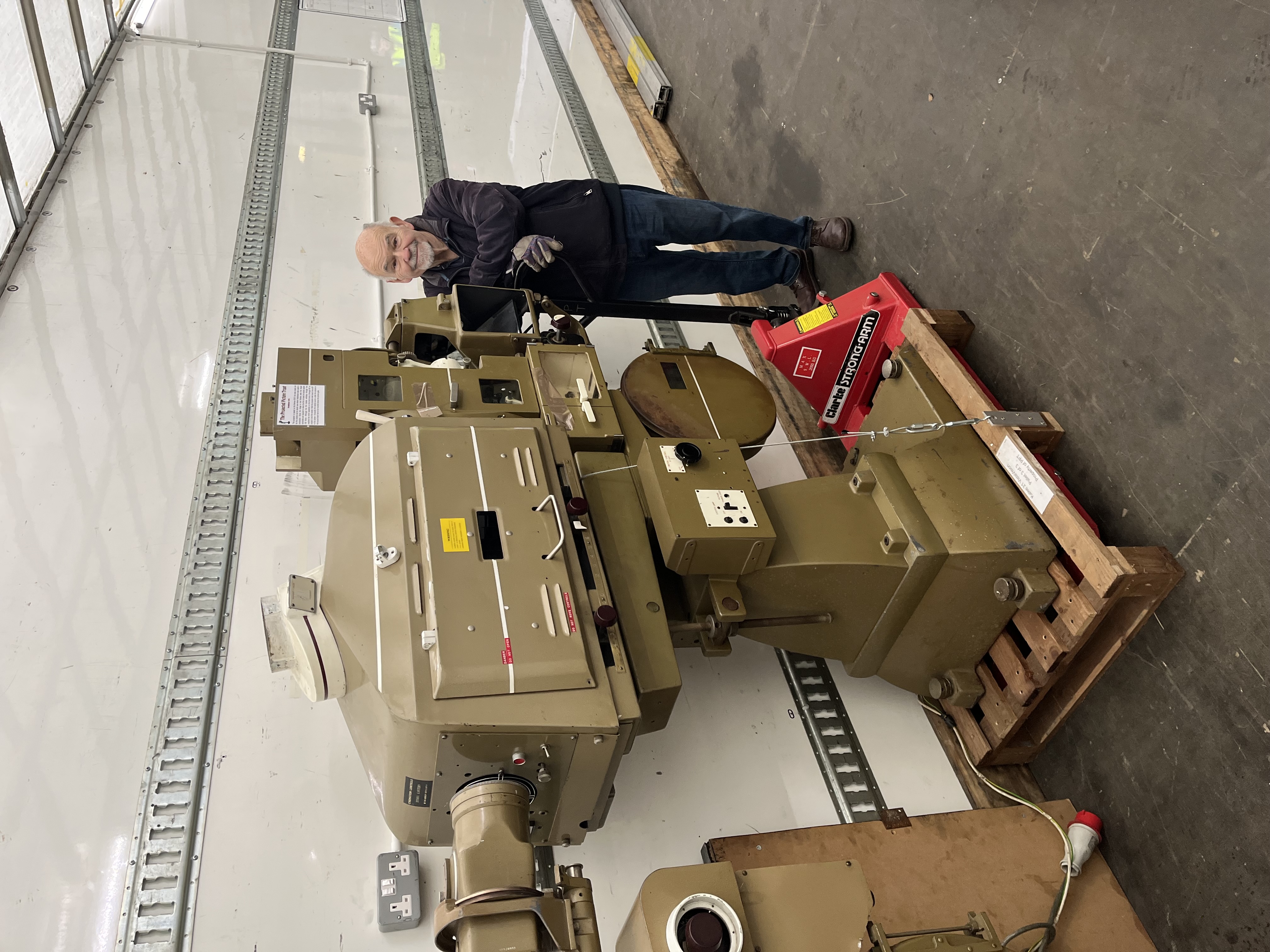 A man standing beside a brown/grey cinema projector. There is a mirror and prism on the front of the projector.