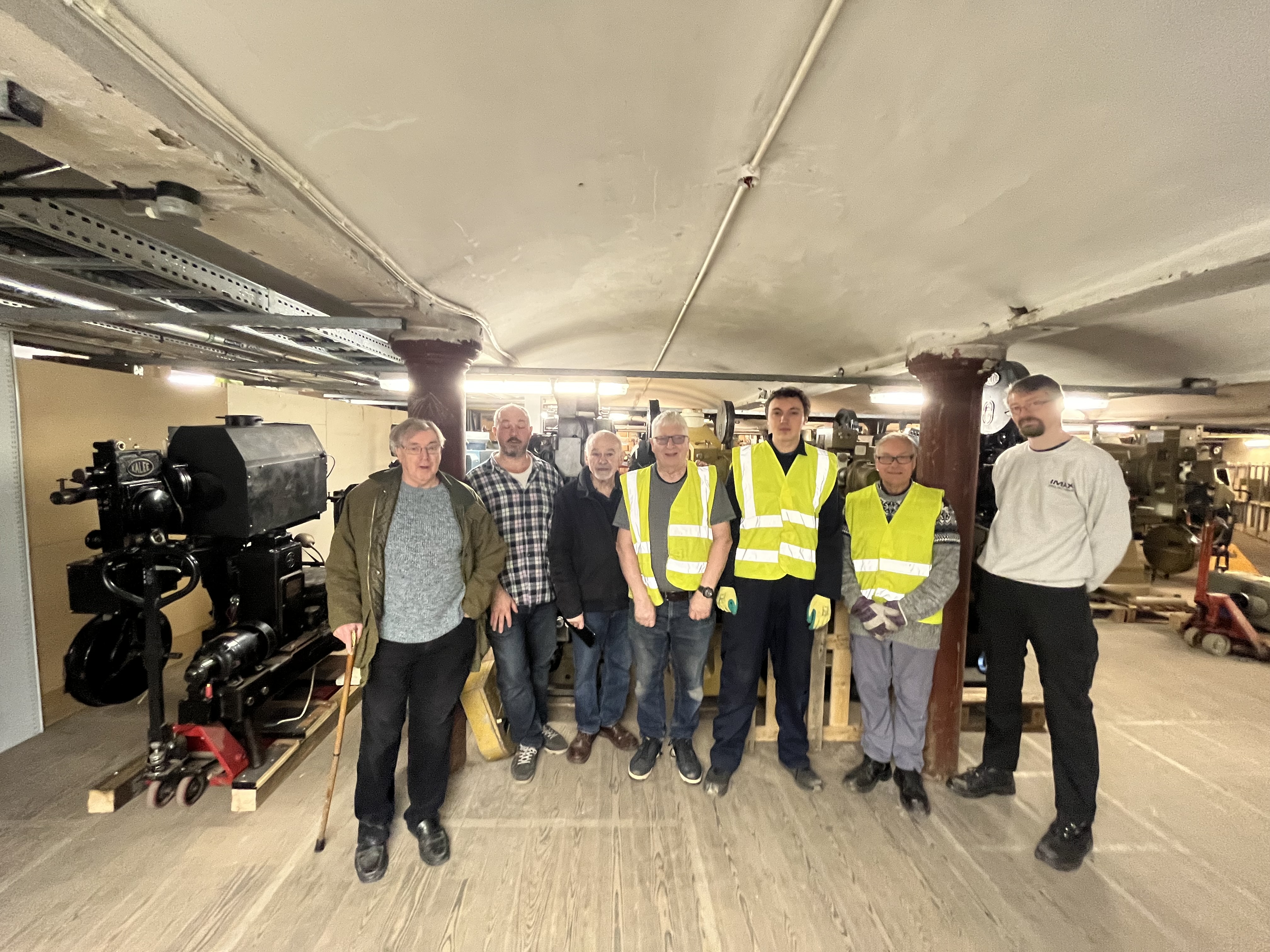Seven men standing in front of cinema projectors and equipment, in a basement.