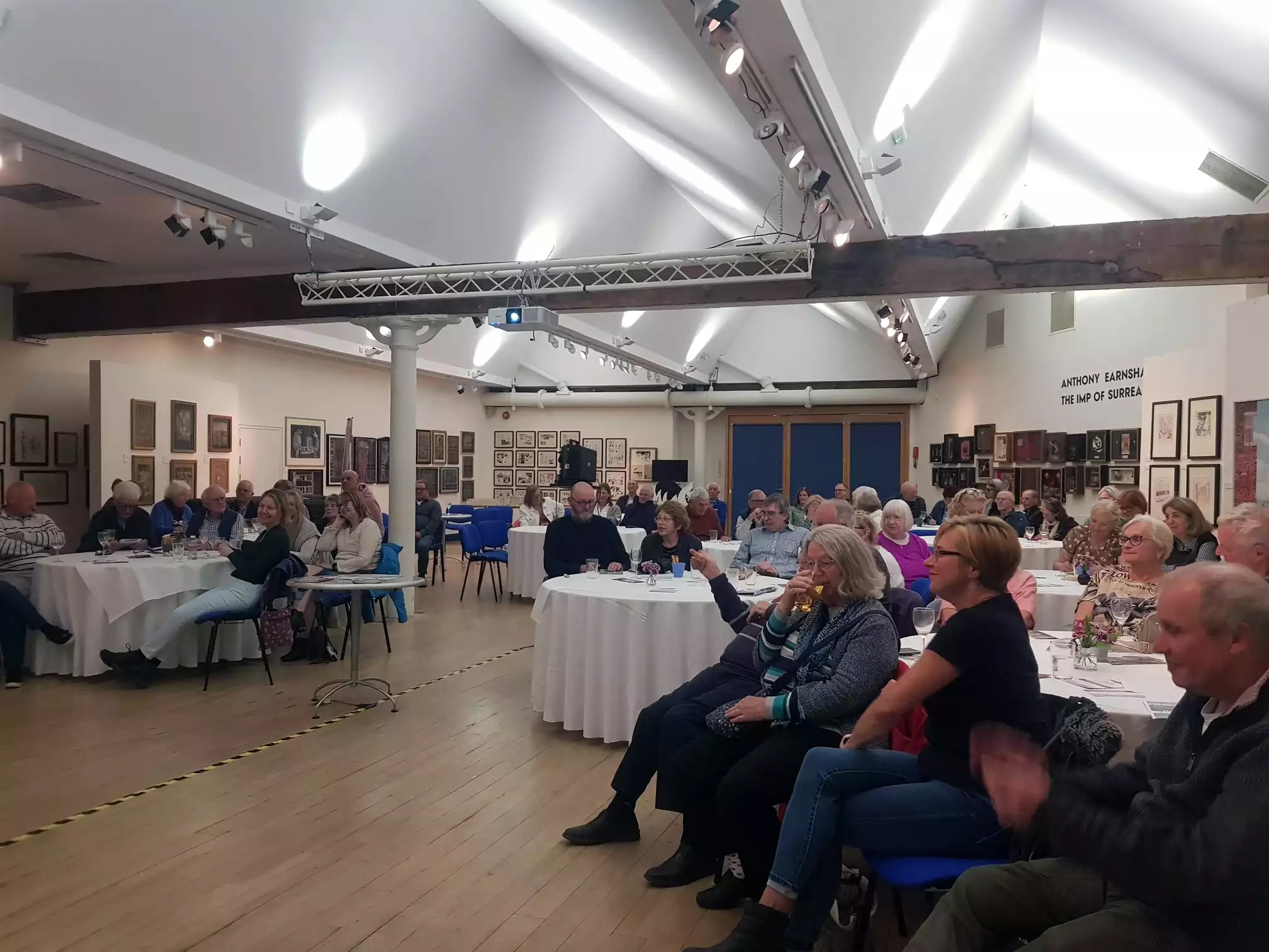 An art gallery with people seated around white tables, looking forward at a screen