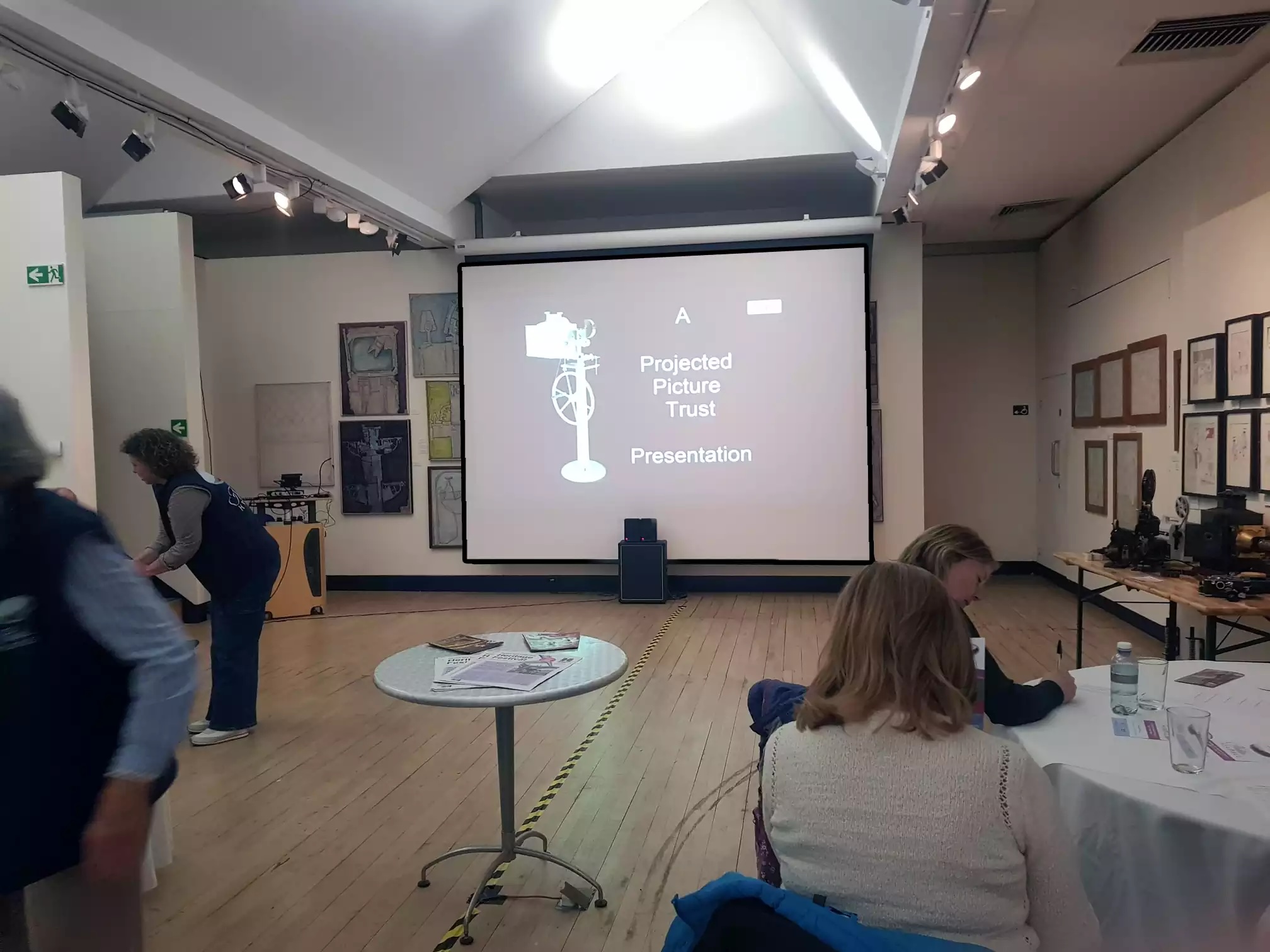 An art gallery, with a projector screen reading "A Projected Picture Trust Presentation"