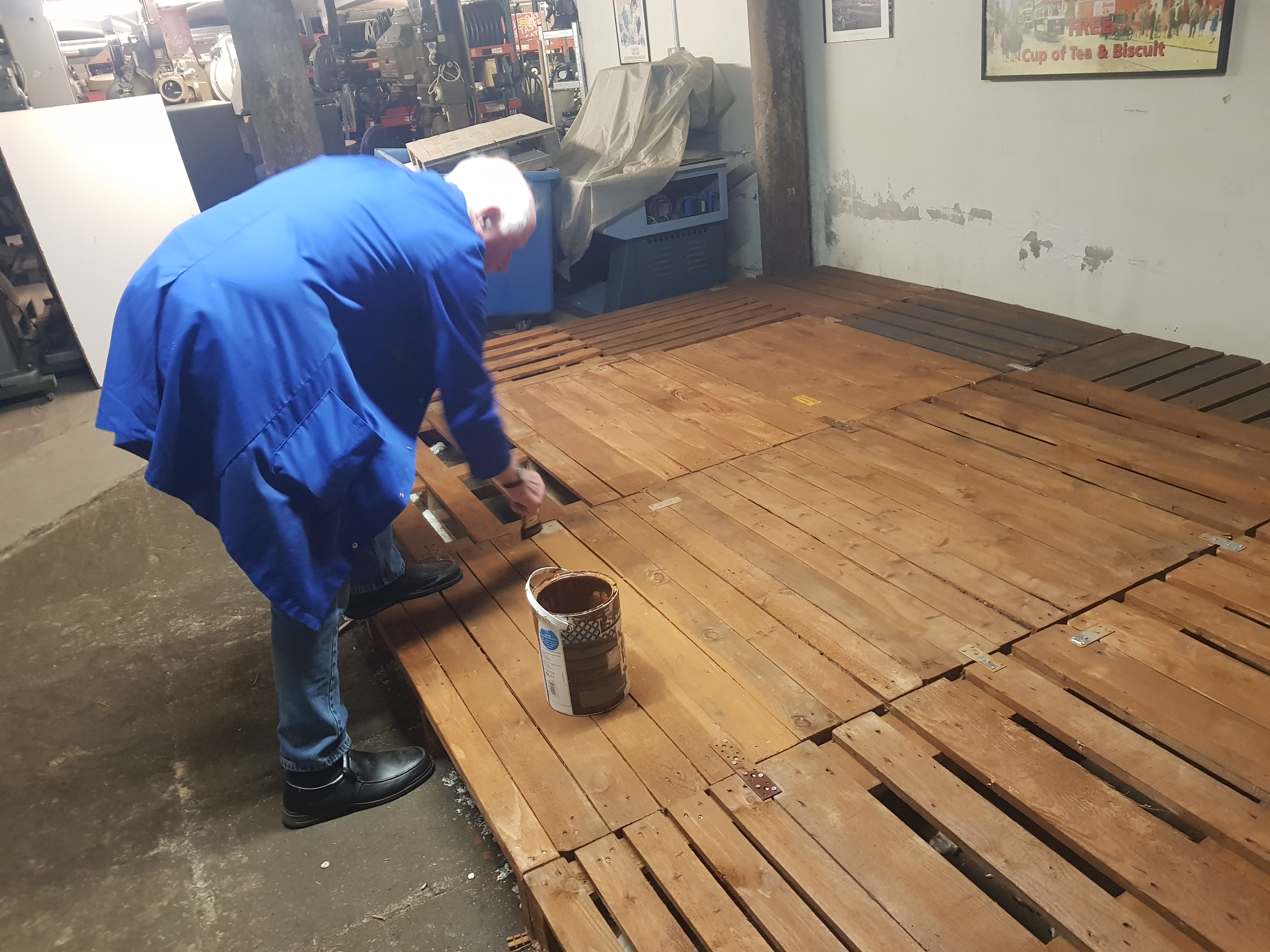 A man in a blue overcoat painting wooden pallets on the floor with brown paint