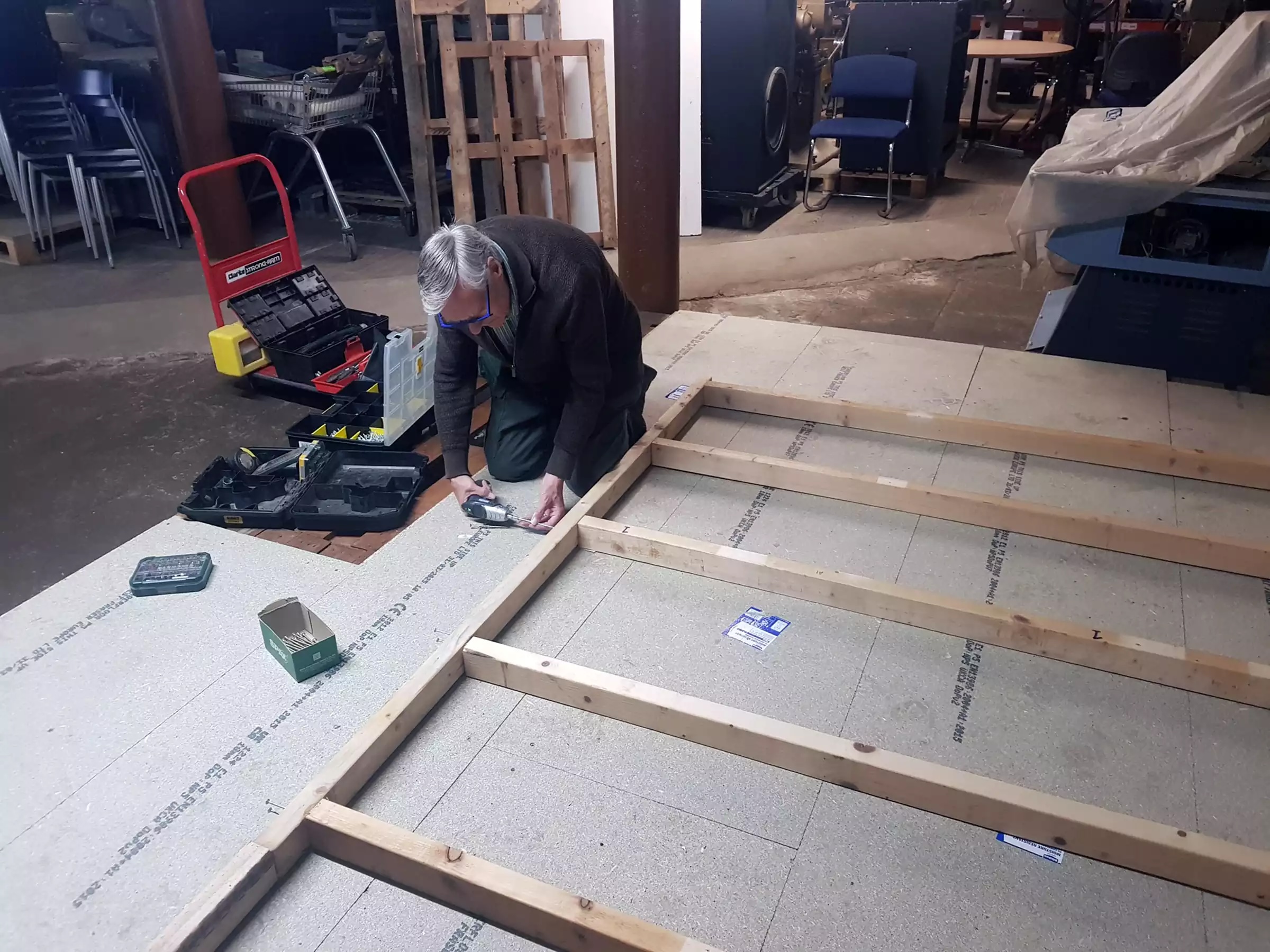 A man kneeling on the floor, using power tools to construct a wooden stud wall